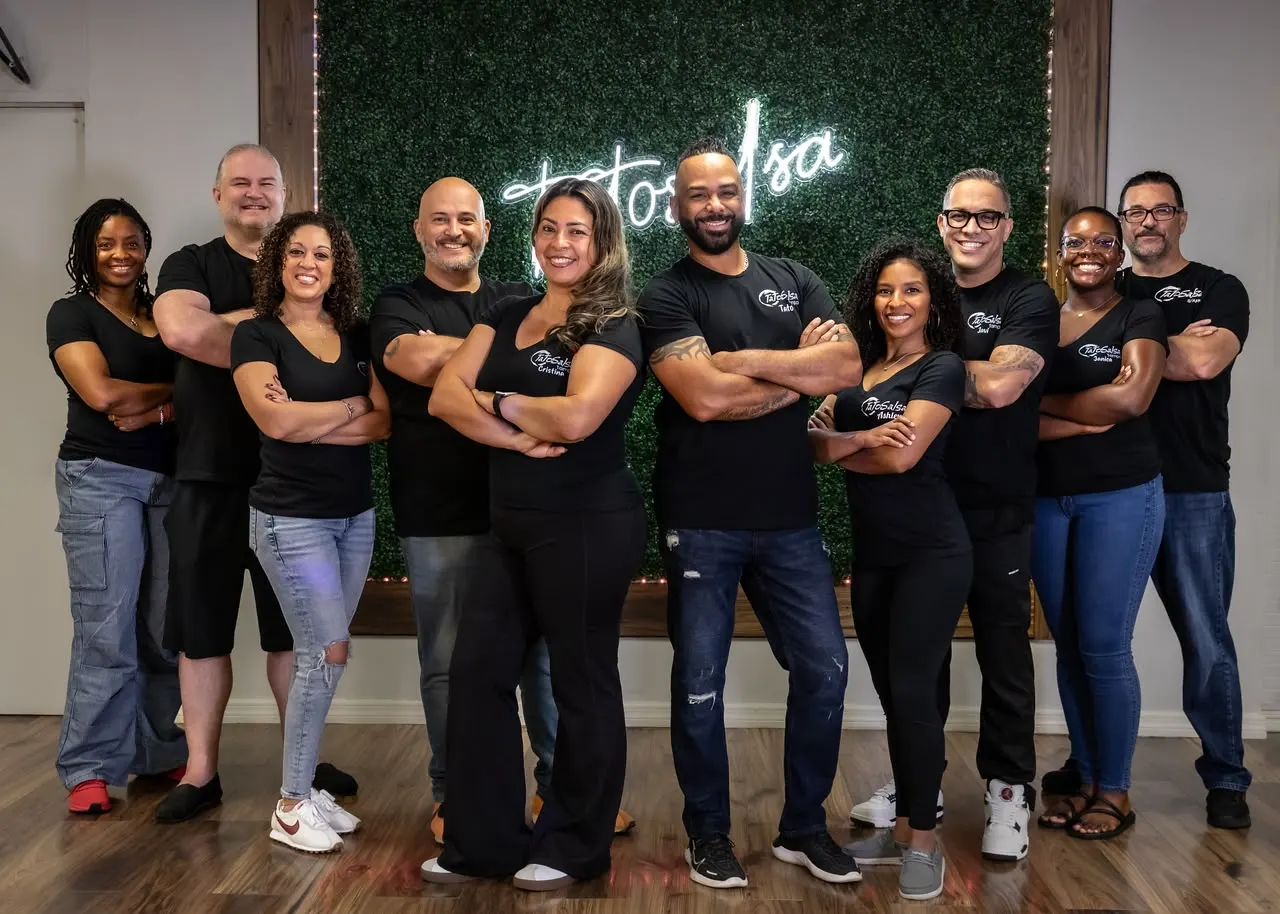 The Tato Salsa staff in black studio shirts, arms crossed, in front of the studio's neon sign.